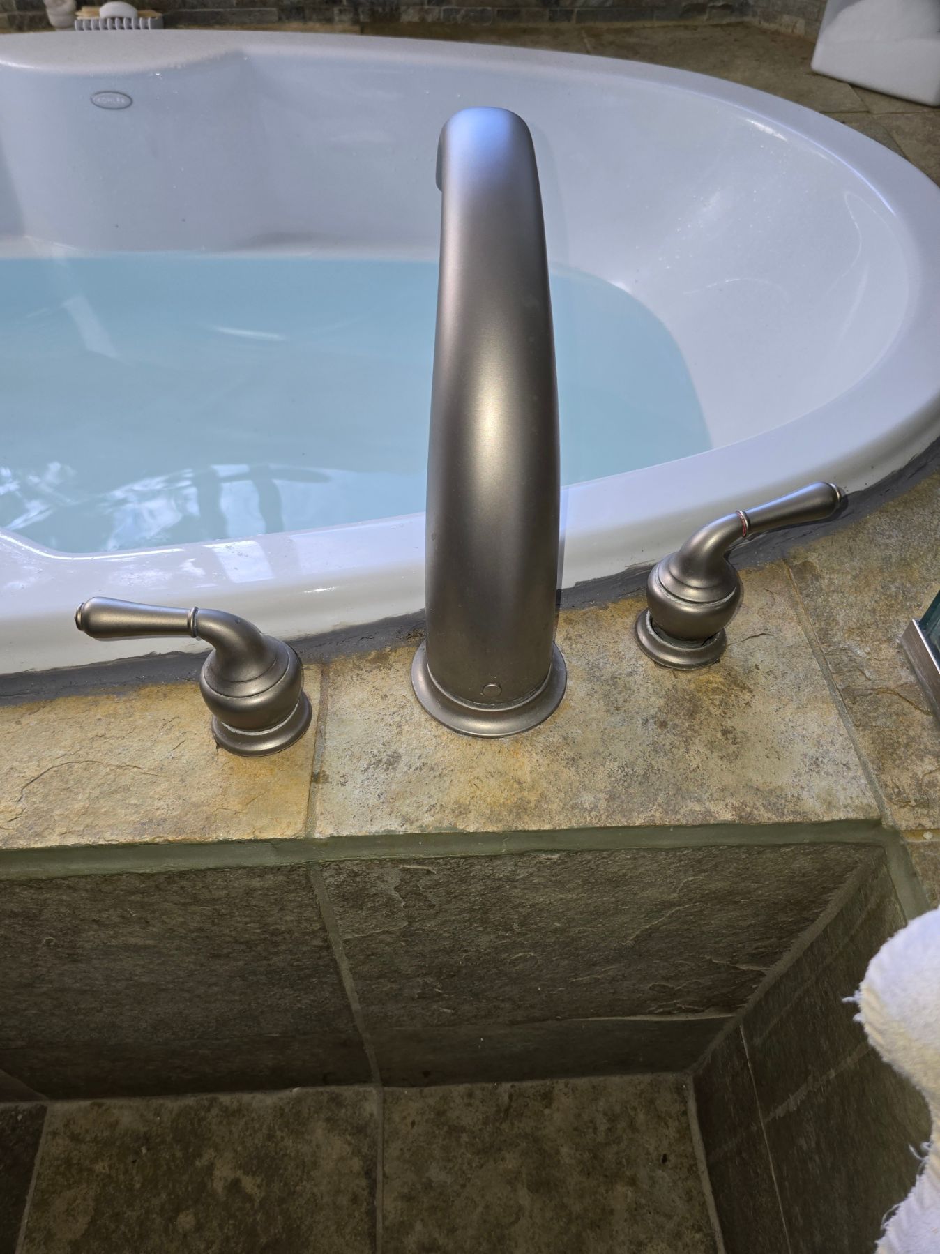 Built-in bathtub with water and three deck-mounted faucets in a tiled bathroom.