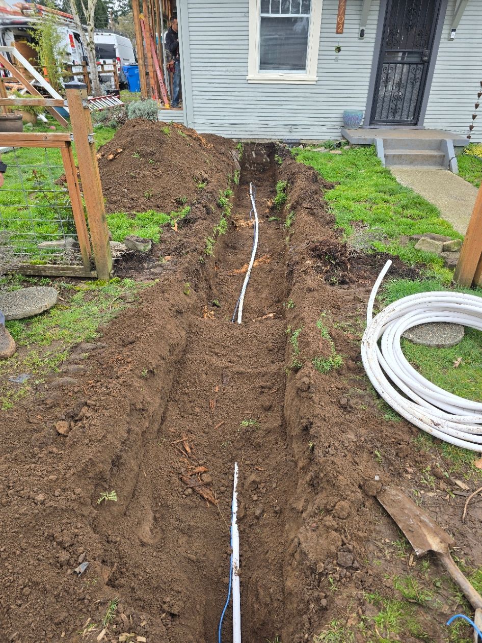 Trench dug in muddy yard beside a house, with a white pipe running through it and coiled pipe nearby.