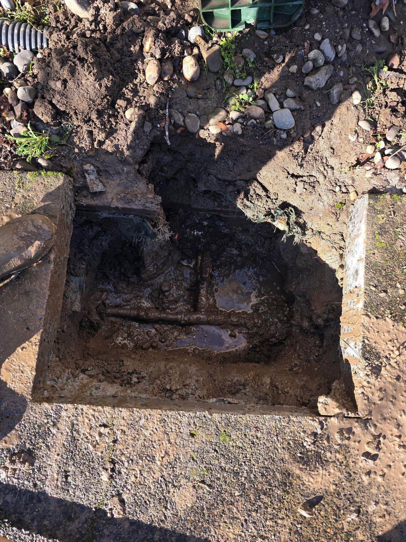 An excavated dirt hole with a dark rectangular opening beside a rough gravel and soil surface.