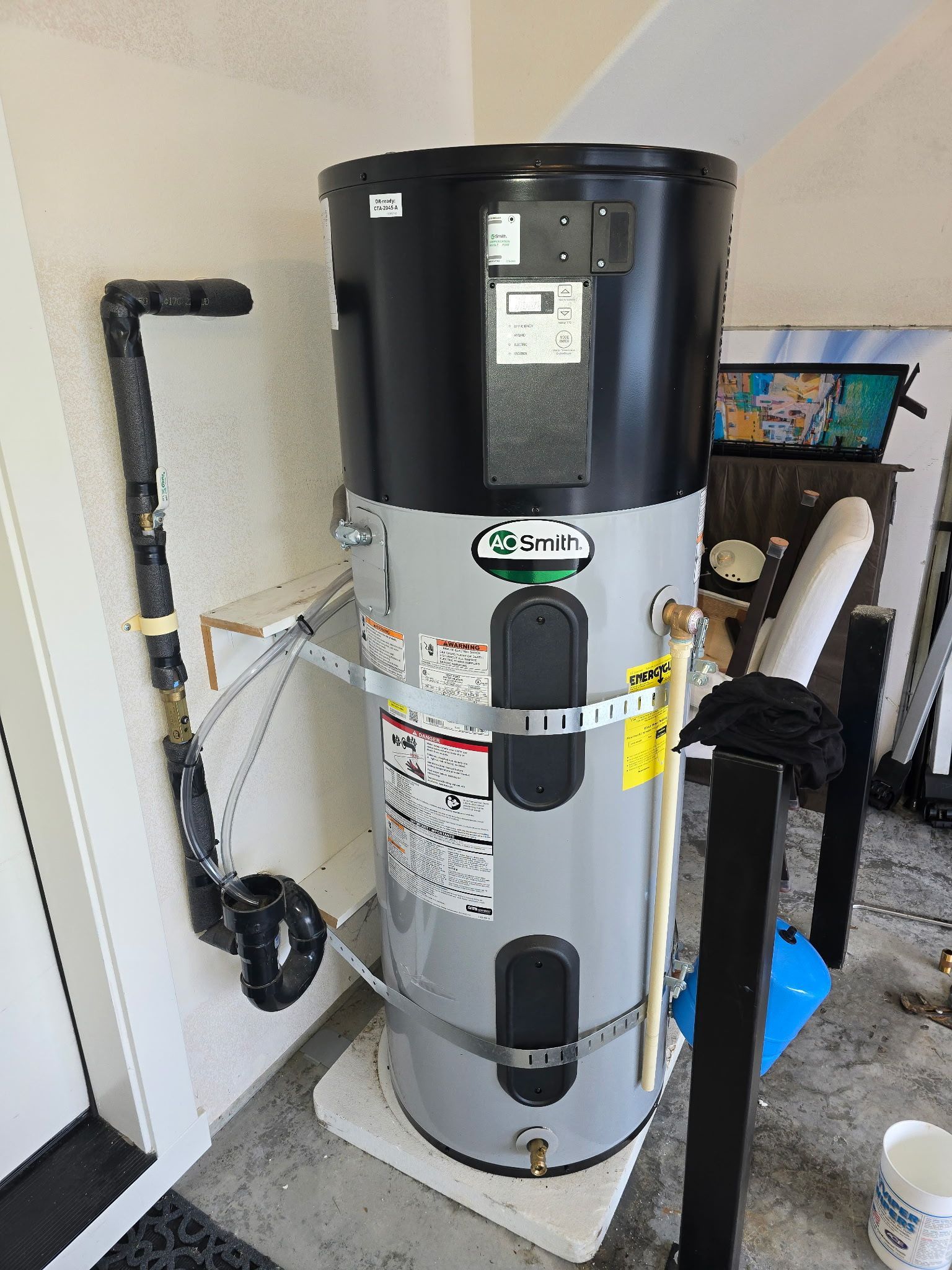 Tall water heater tank in a utility room beside a wall and cords, with a black top and gray body.