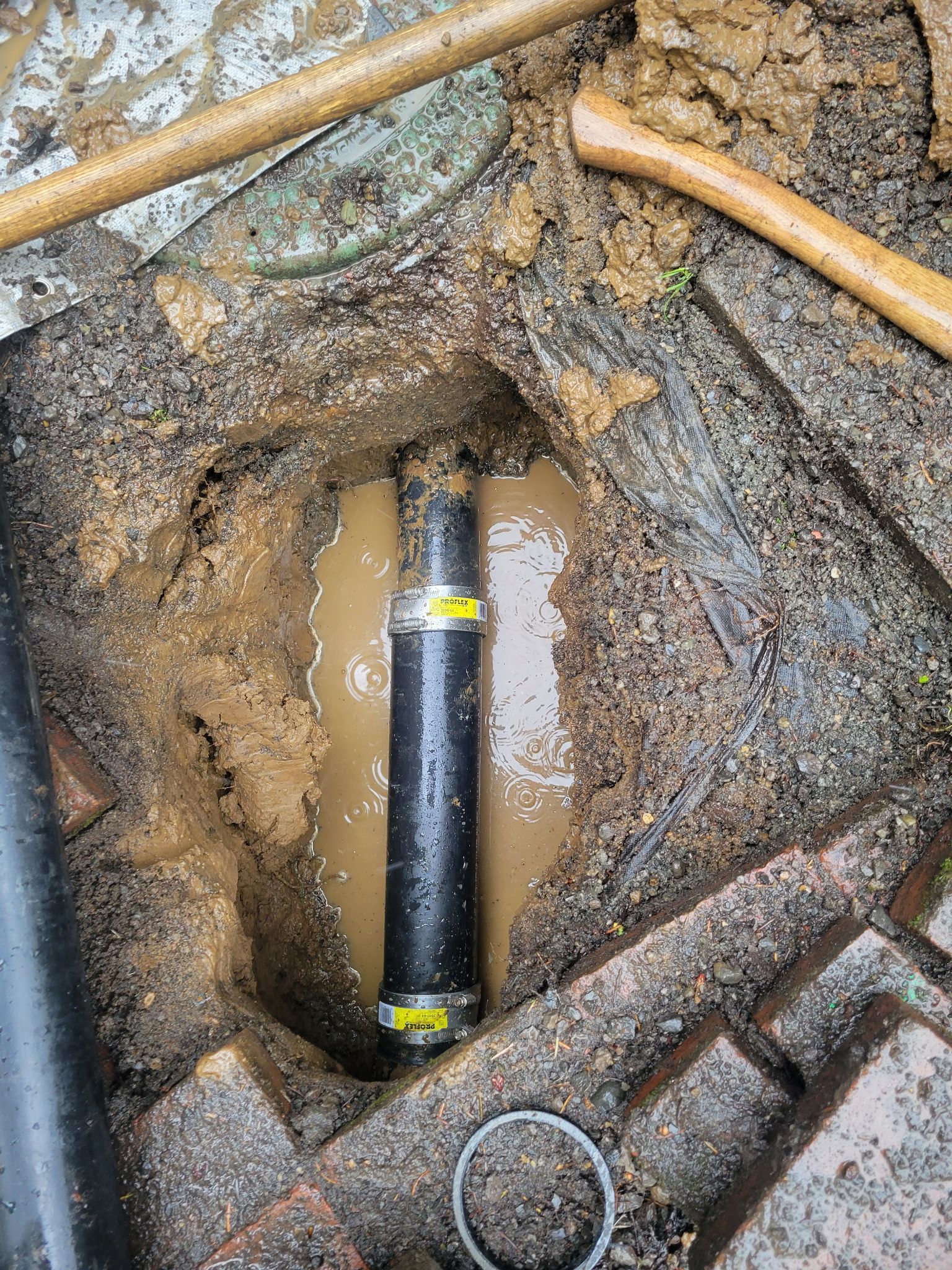 Excavated trench with muddy water and a black underground pipe connection exposed.