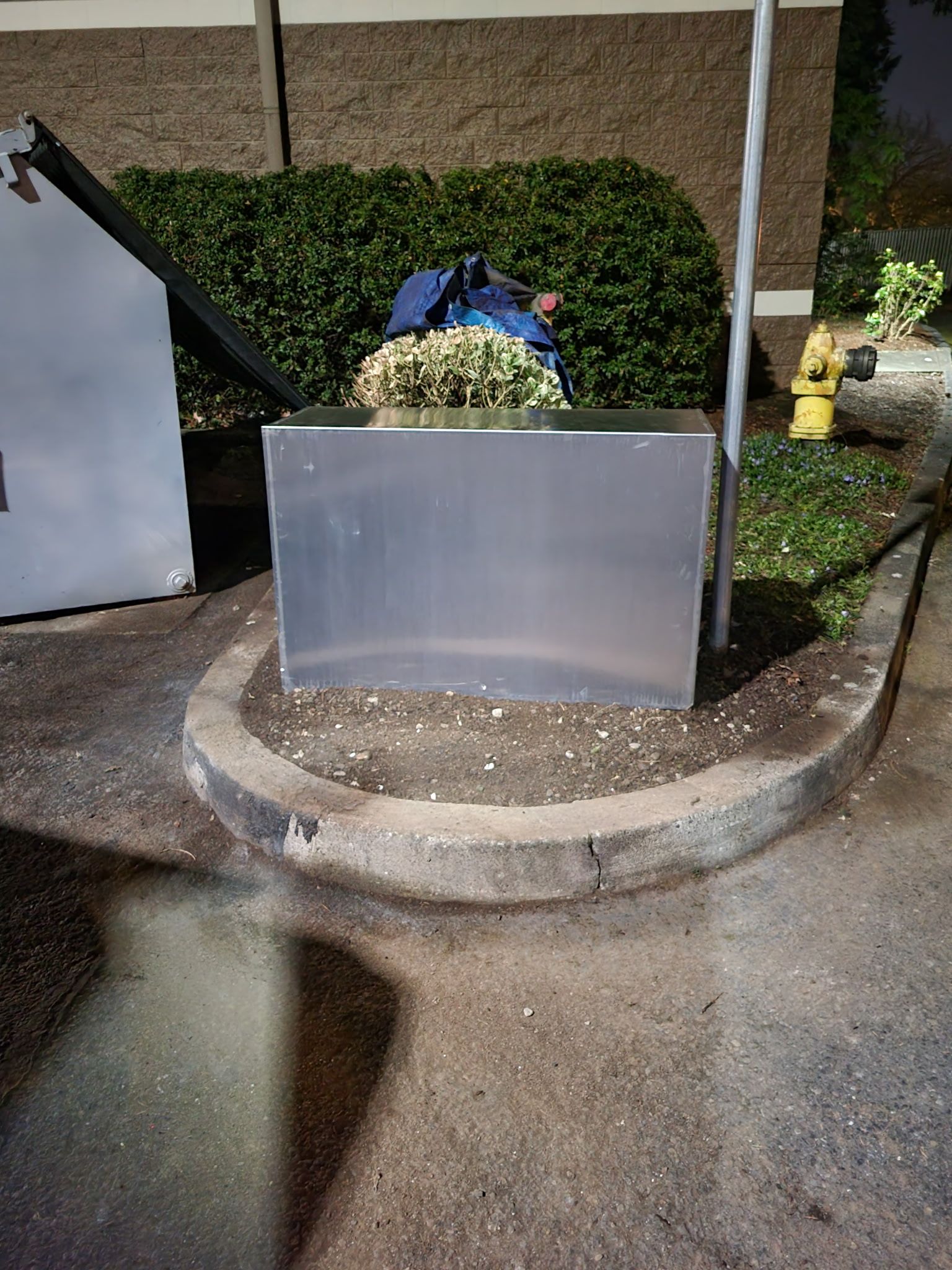 Dustpan and debris container beside a hedge in a wet outdoor area at night