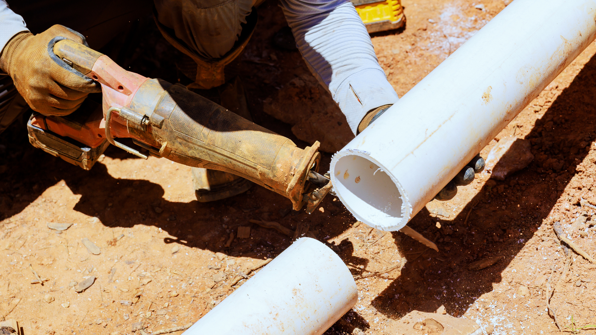 Worker cutting a white PVC pipe with a saw on a dirt construction site