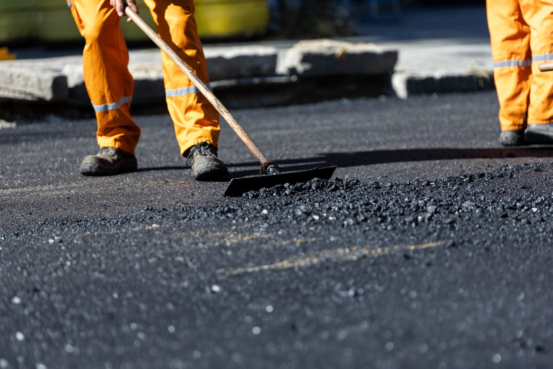 Photograph showing two construction workers in orange safety pants paving a road with fresh asphalt.