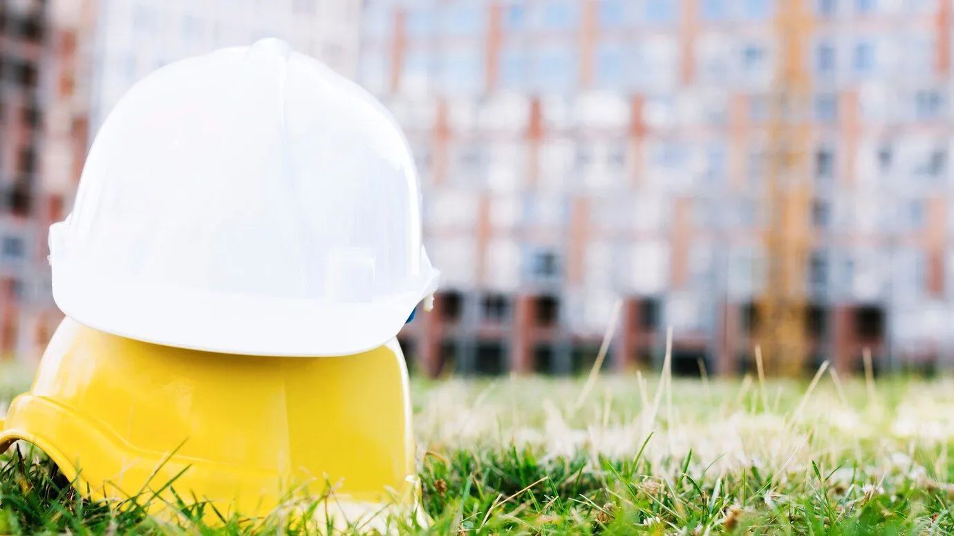 Two construction helmets, yellow and white, stacked on grass with a city highrise in the background