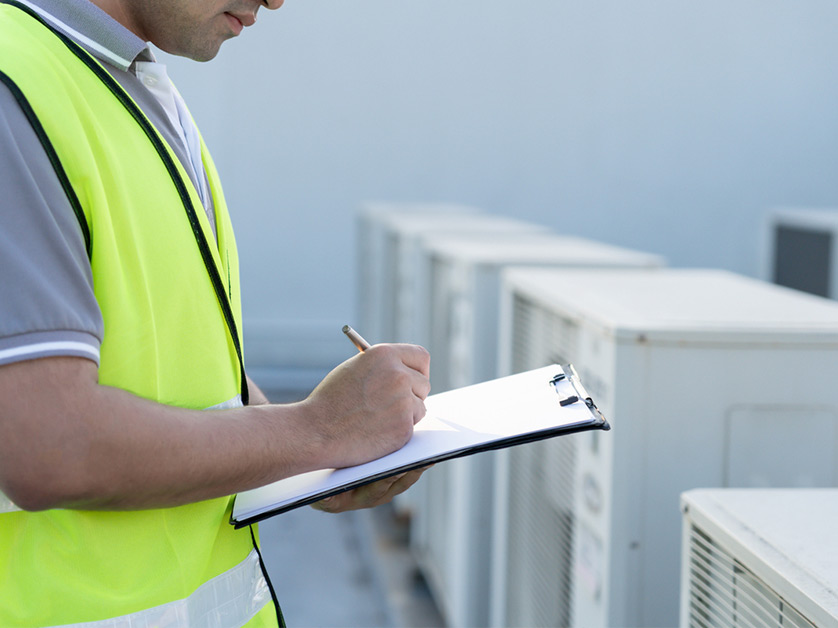 HVAC technician in a safety vest inspecting and documenting air conditioning units on a rooftop.