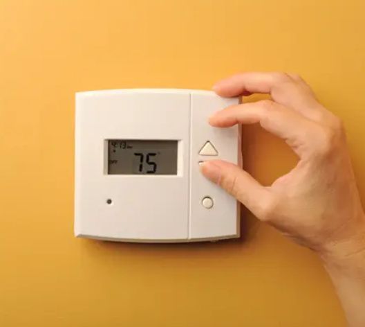 Hand adjusting a white thermostat on a yellow wall, setting the temperature to 75.