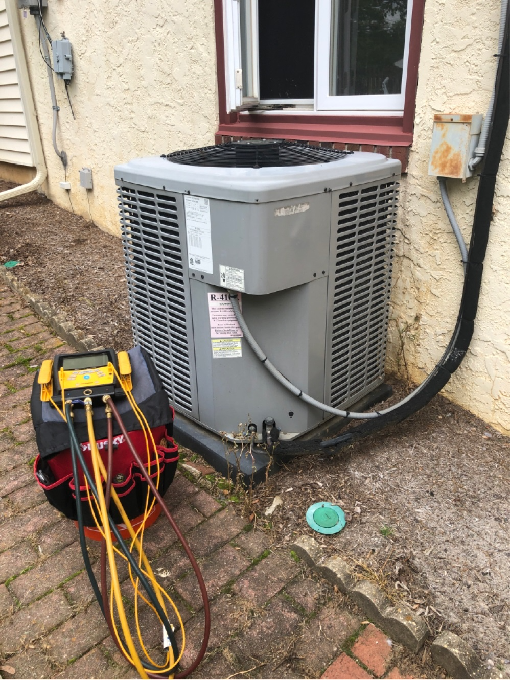 HVAC unit with attached gauges. Outdoors, brick patio. Grey and yellow equipment. Black hose.