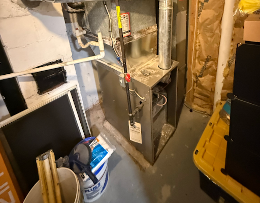 Furnace in a basement; metal, rectangular, pipes attached. There is a bucket and other items in the foreground.