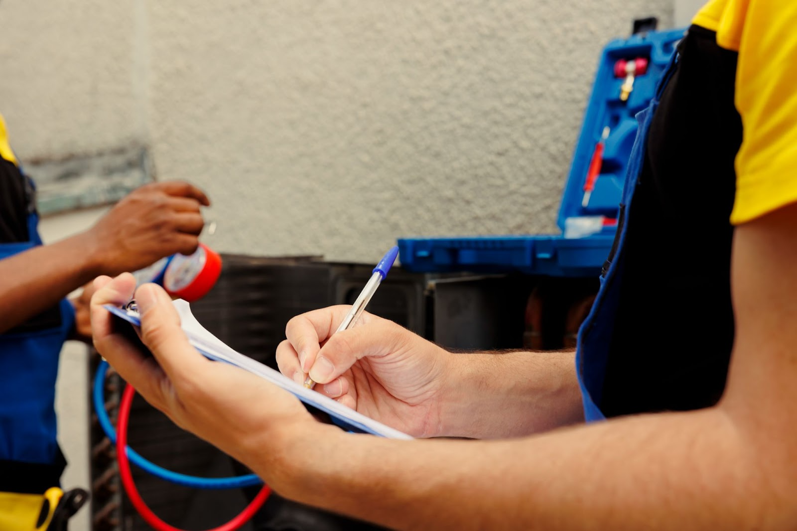 HVAC technician writing on clipboard, another works on equipment, blue toolbox visible.