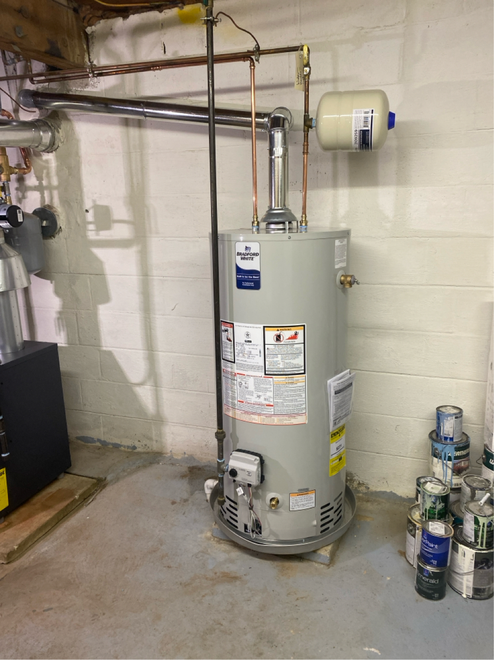 Water heater in a basement against a white cinder block wall with copper pipes and a small expansion tank.