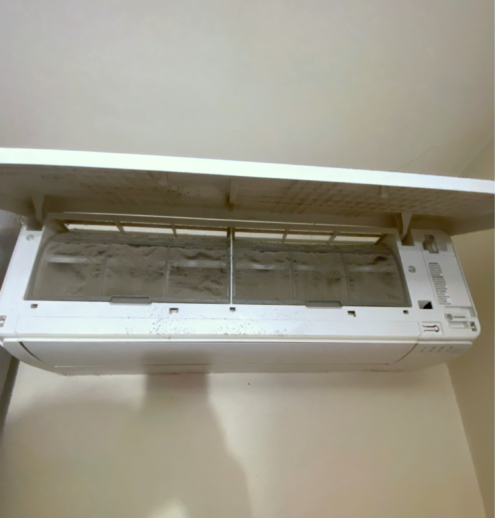 A dirty, white air conditioner with an open vent, covered in dust. Mounted on a wall.