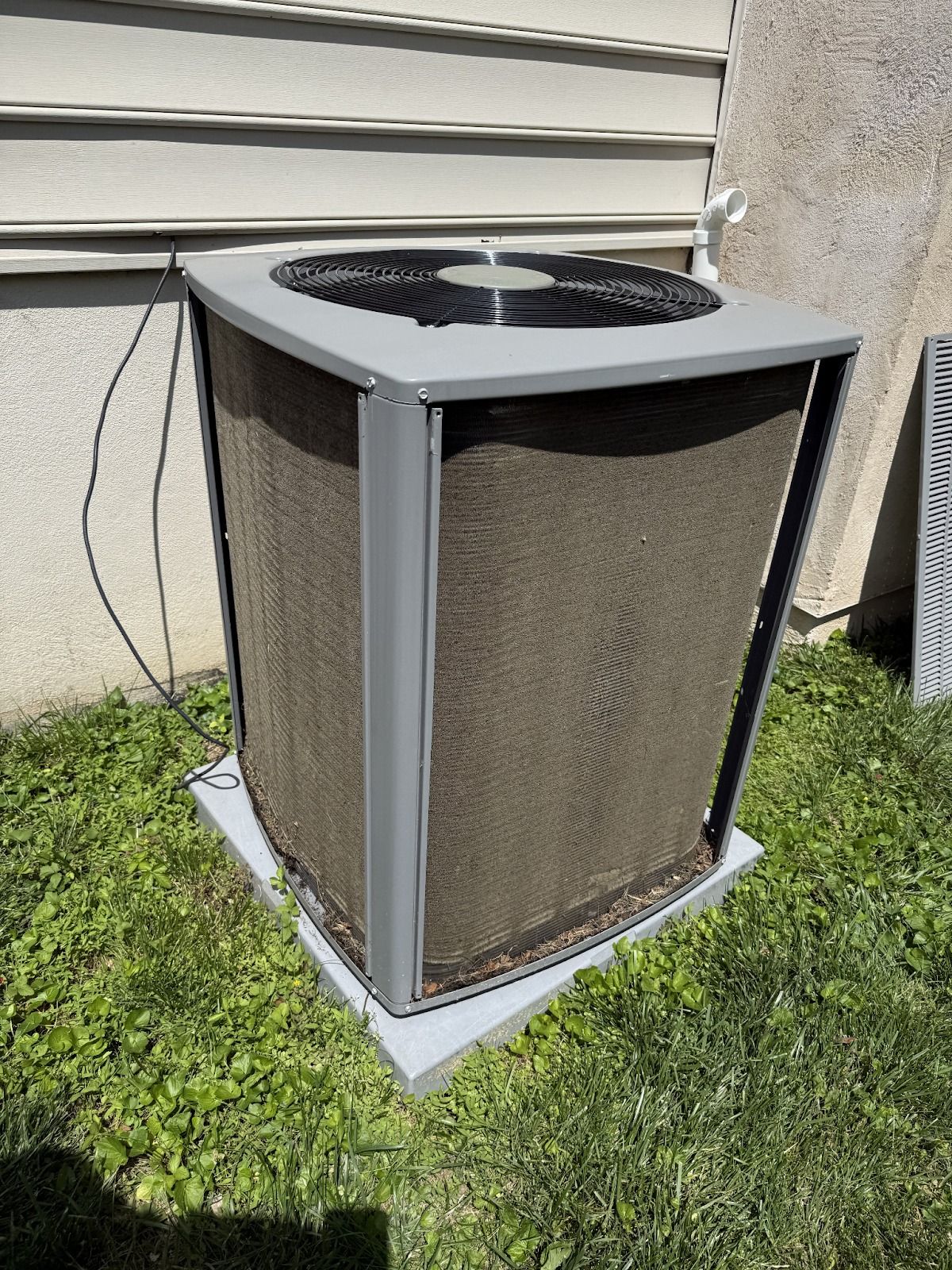 Outdoor air conditioning unit with a dark mesh covering, on a concrete pad, surrounded by grass.