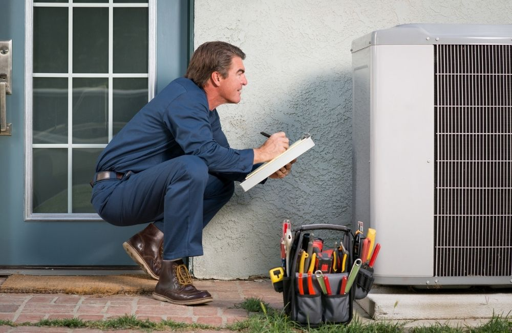 HVAC technician inspecting an air conditioning unit outside a building, writing on a clipboard.