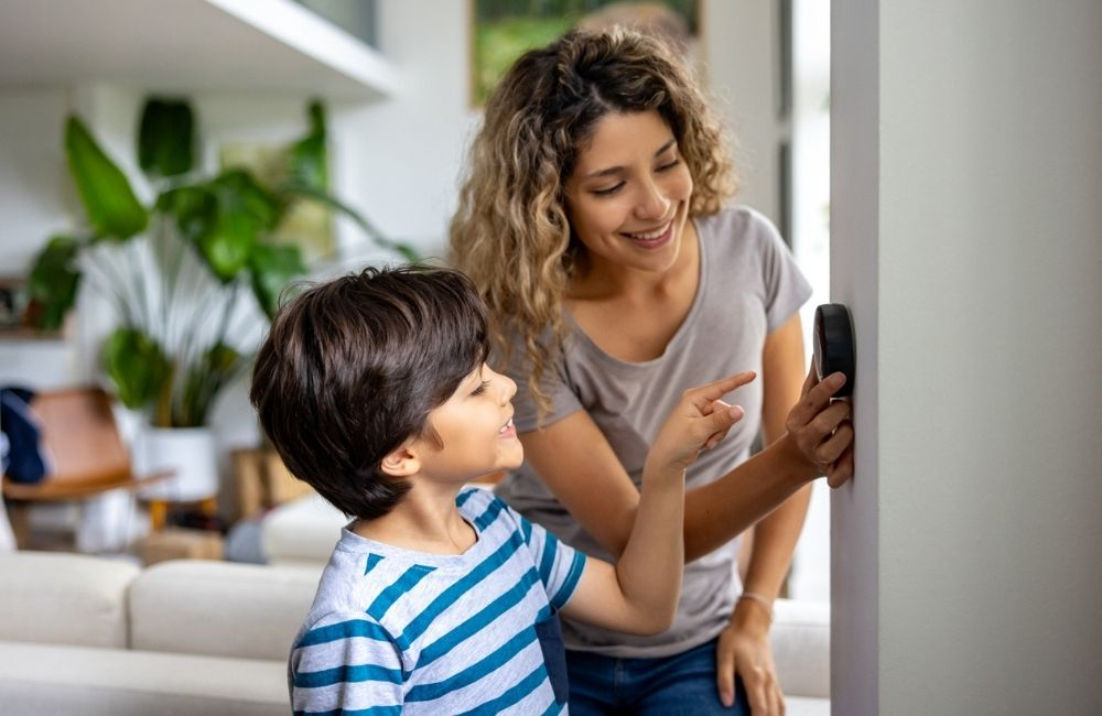Woman and child pointing at a wall-mounted smart device indoors; child's finger touching device.