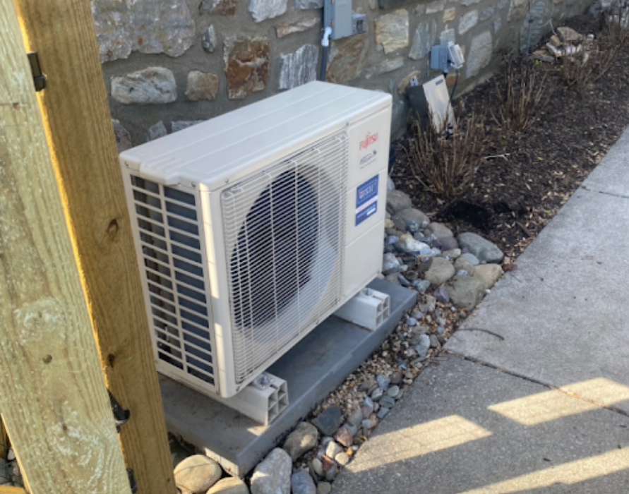 Fujitsu heat pump unit next to a stone wall, on concrete base, with rocks and sidewalk.