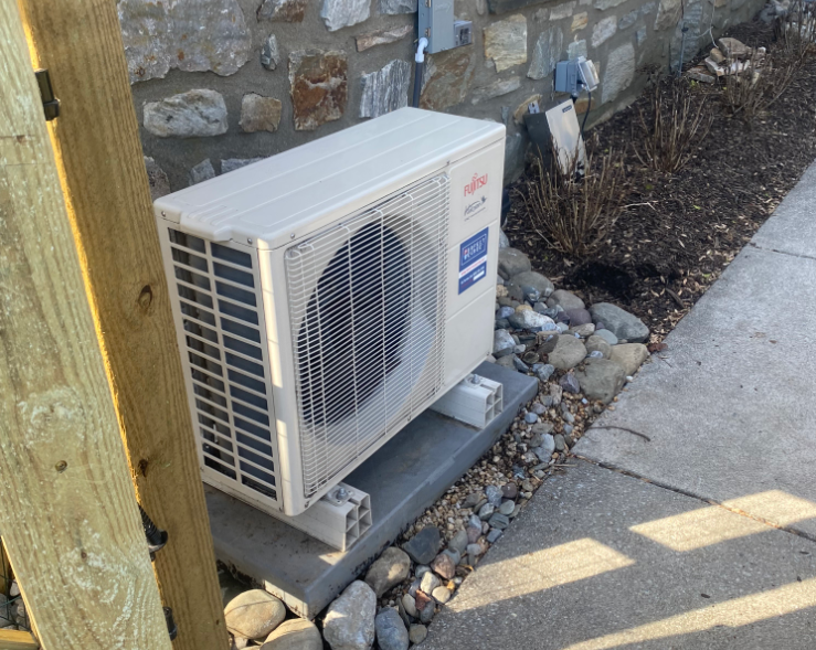 A white Fujitsu outdoor HVAC unit sits on a concrete pad with cinder block risers, located against a stone wall.