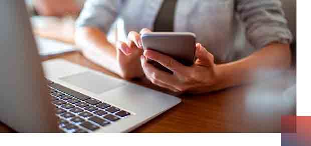 Person using a smartphone and laptop at a desk. Hands hold phone, typing on a laptop.