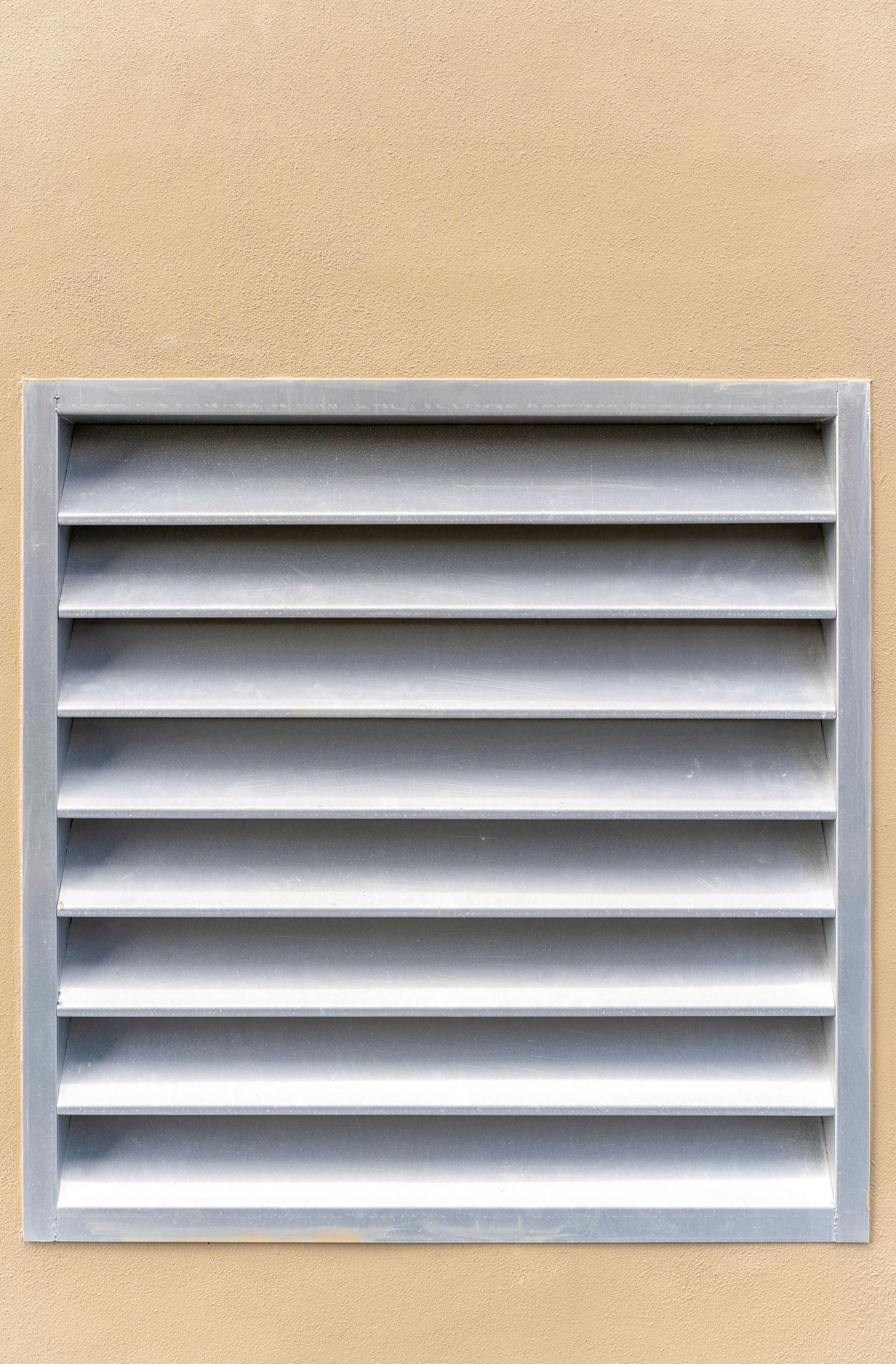 Gray rectangular louvered vent on a beige textured wall.