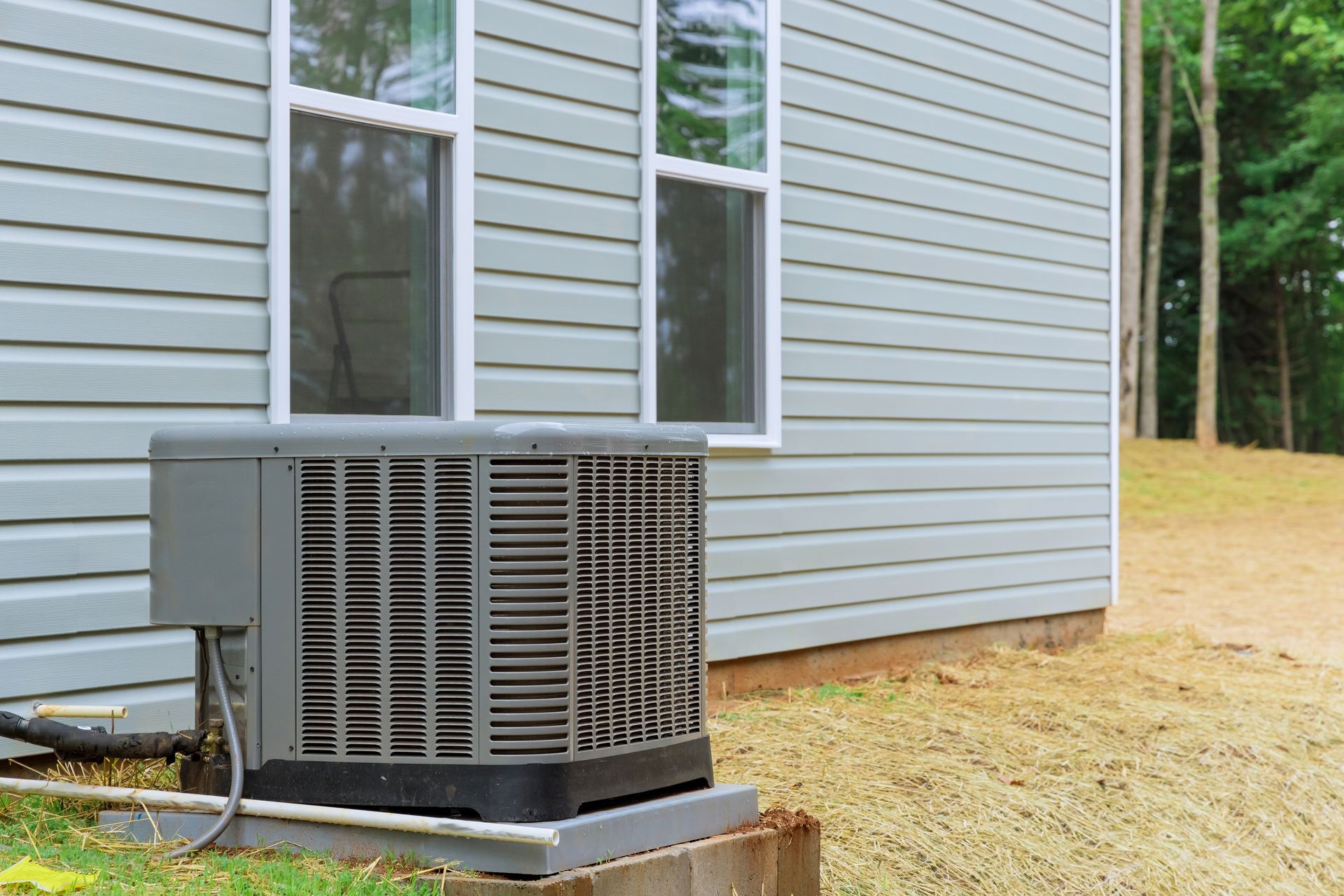 A gray outdoor HVAC unit sits on a concrete pad next to a house with light gray vinyl siding and two windows.
