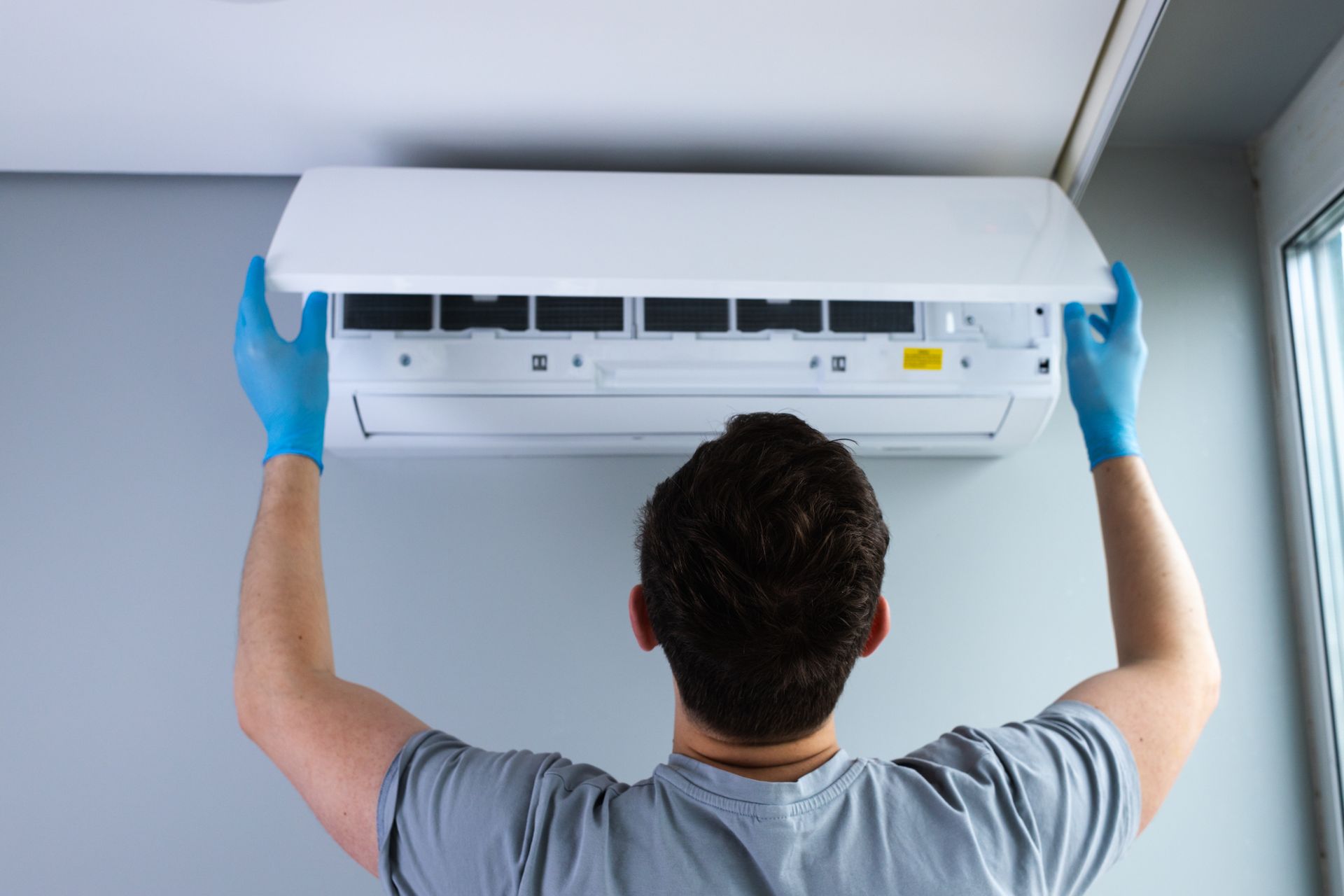 Person in gloves opening an air conditioner, mounted on a gray wall.