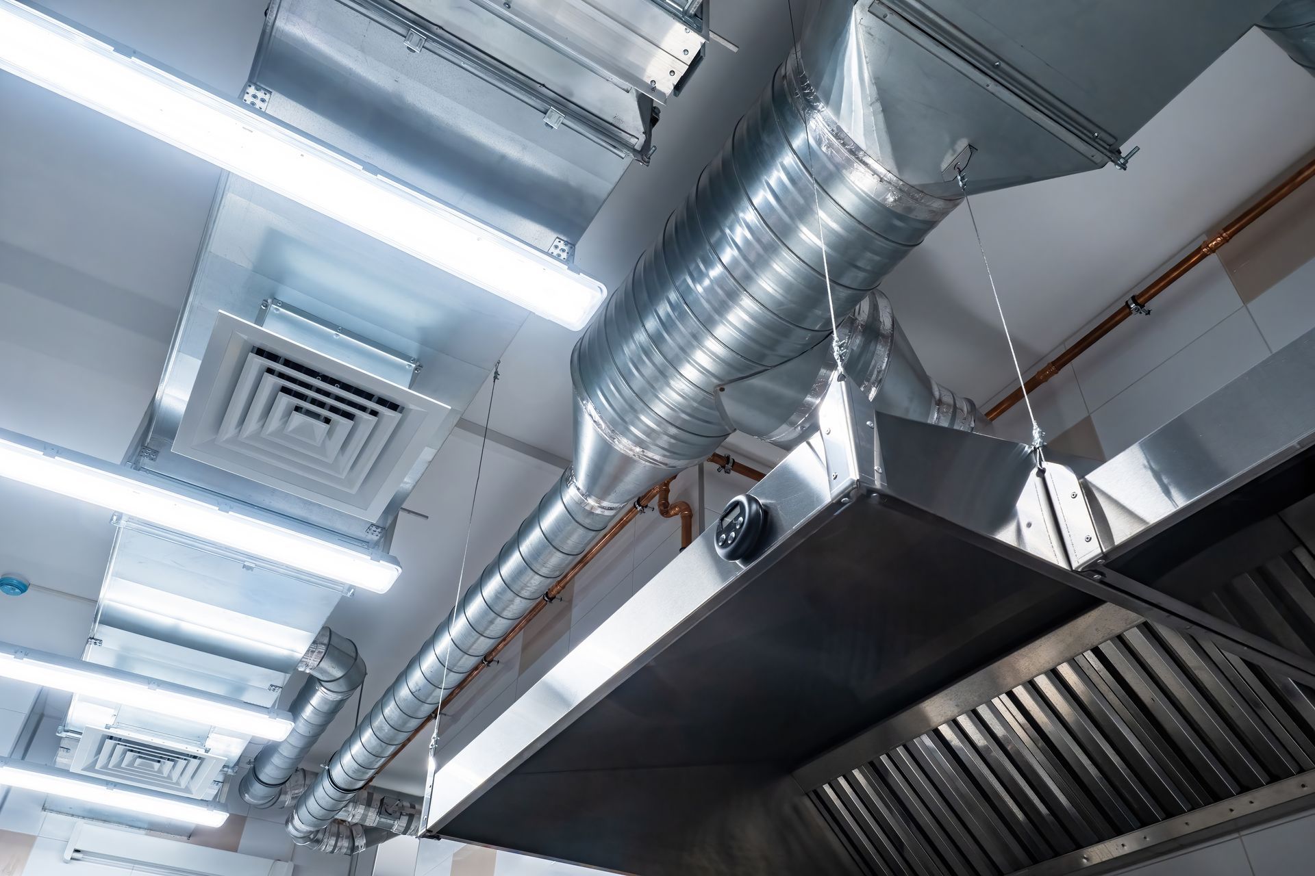 A close-up of a stainless steel kitchen ventilation hood connected to a round industrial duct system under a lit ceiling.