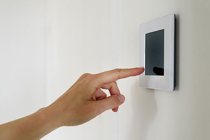 Hand pointing at a rectangular, dark-screened device on a white wall.
