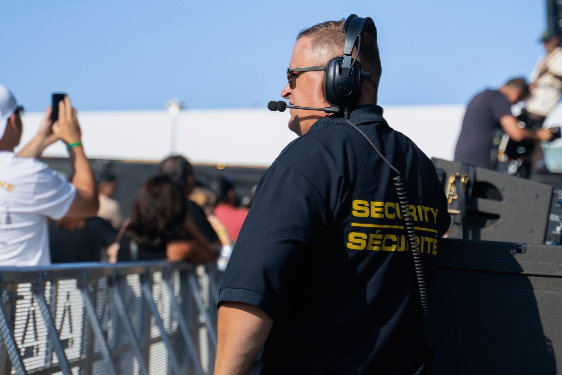 A security guard wearing headphones and a microphone is standing in front of a crowd of people.