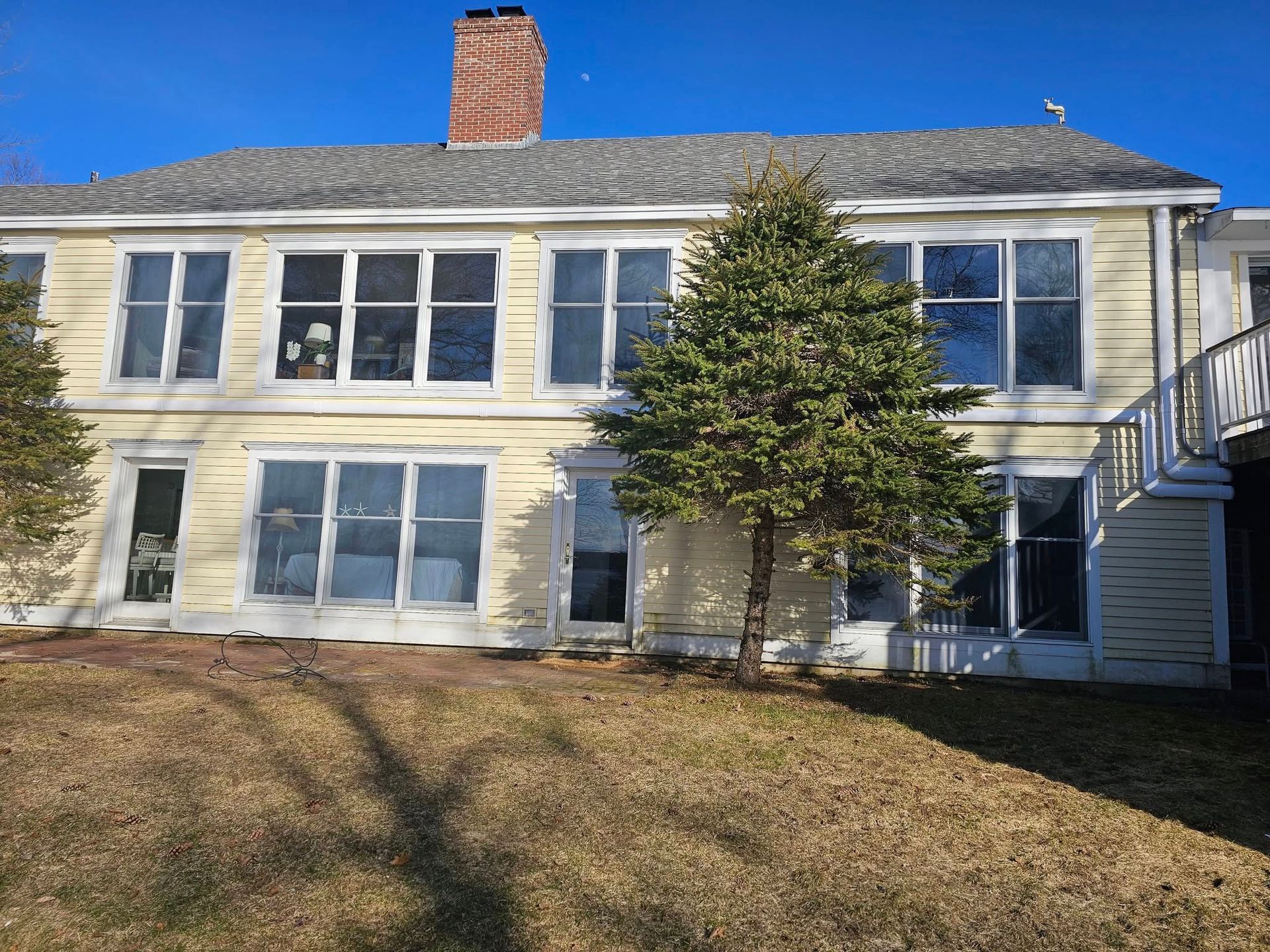 Two-story yellow house with multiple windows, a chimney, and a small tree in front on a sunny day.