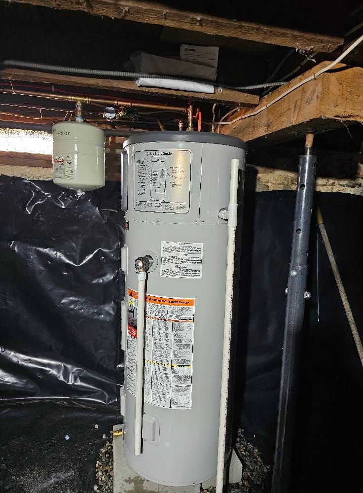 Water heater in a basement. Grey tank, white expansion tank, and pipes against a black plastic-covered wall.