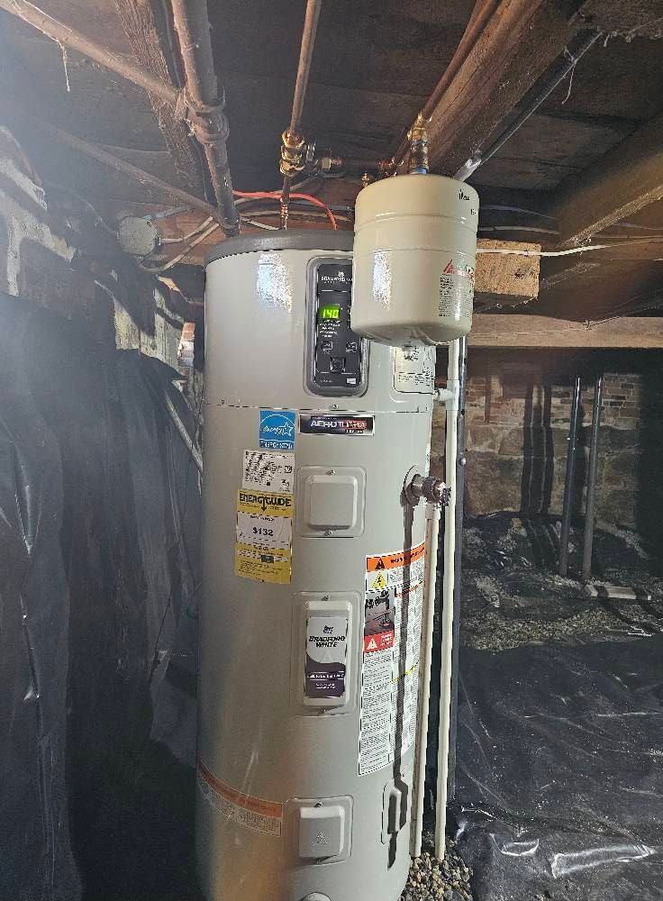 Water heater in a basement. The appliance is tall, cylindrical, and metallic. It has a booster tank on top and several control panels.
