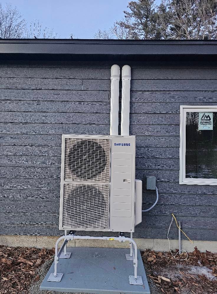 Outdoor heat pump unit mounted on a concrete pad, against a gray building with two white vent pipes.