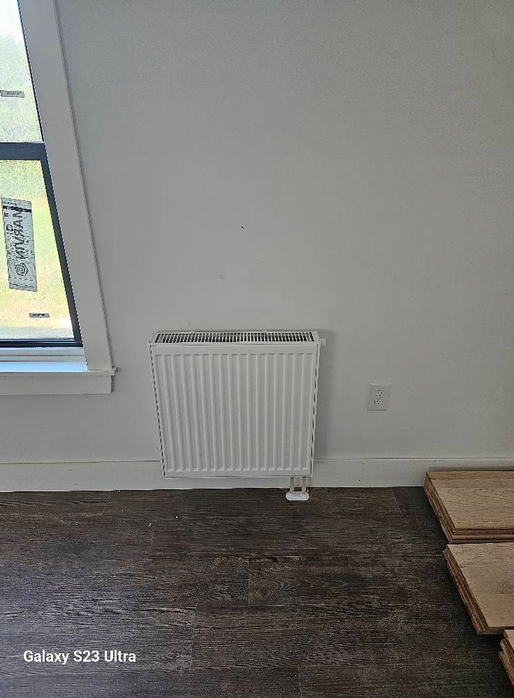 White radiator on a wall, near a window and electrical outlet, with dark wood flooring.