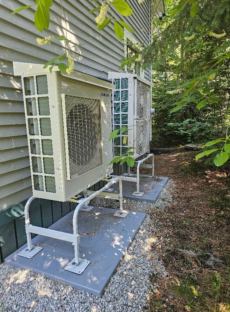 Two outdoor air conditioning units mounted on gray platforms next to a wood-sided building.