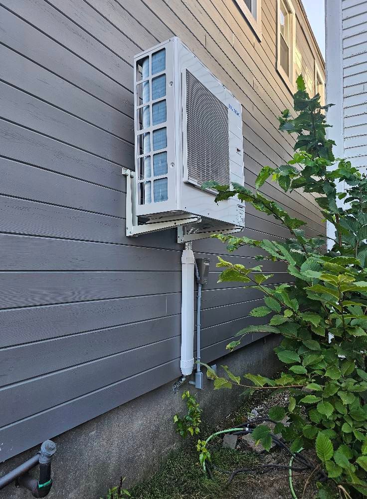 An air conditioning unit mounted on a gray house exterior, with white pipes and green foliage.