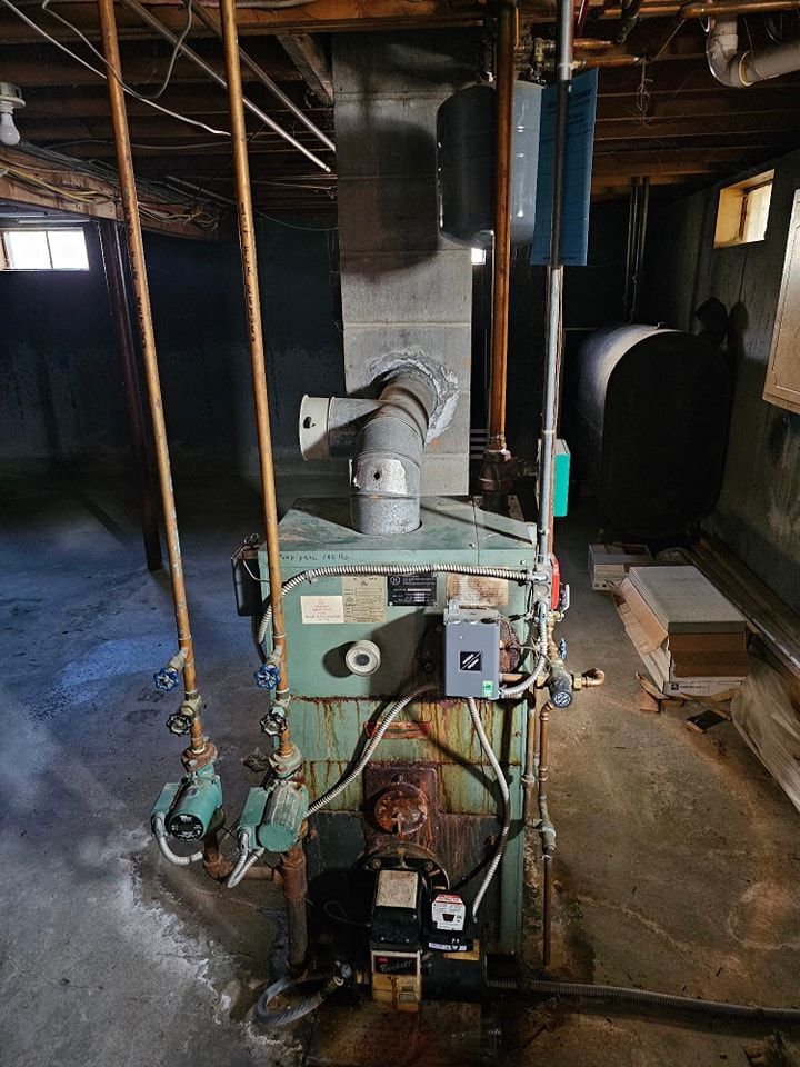 Old, green oil-burning furnace in a basement. Ductwork and pipes visible. A large black oil tank is in the background.