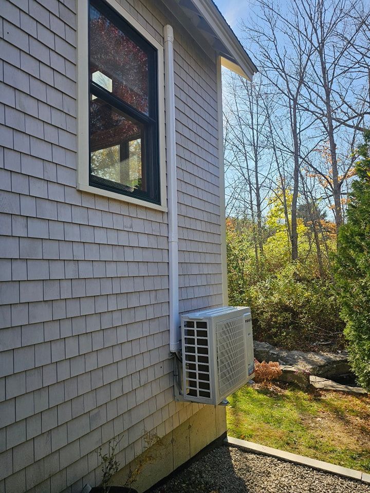 Air conditioner unit mounted on a gray shingled house, white pipe runs up the side near a window, sunny outdoors.
