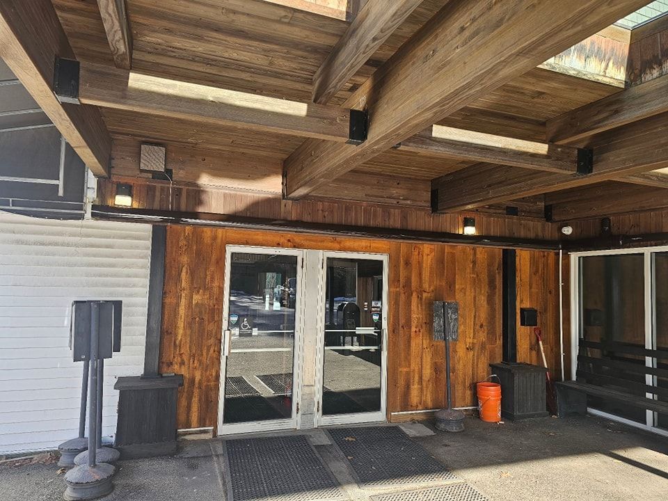 Wooden entrance with glass doors under a wood beam overhang.  Two black trash cans flank the door.