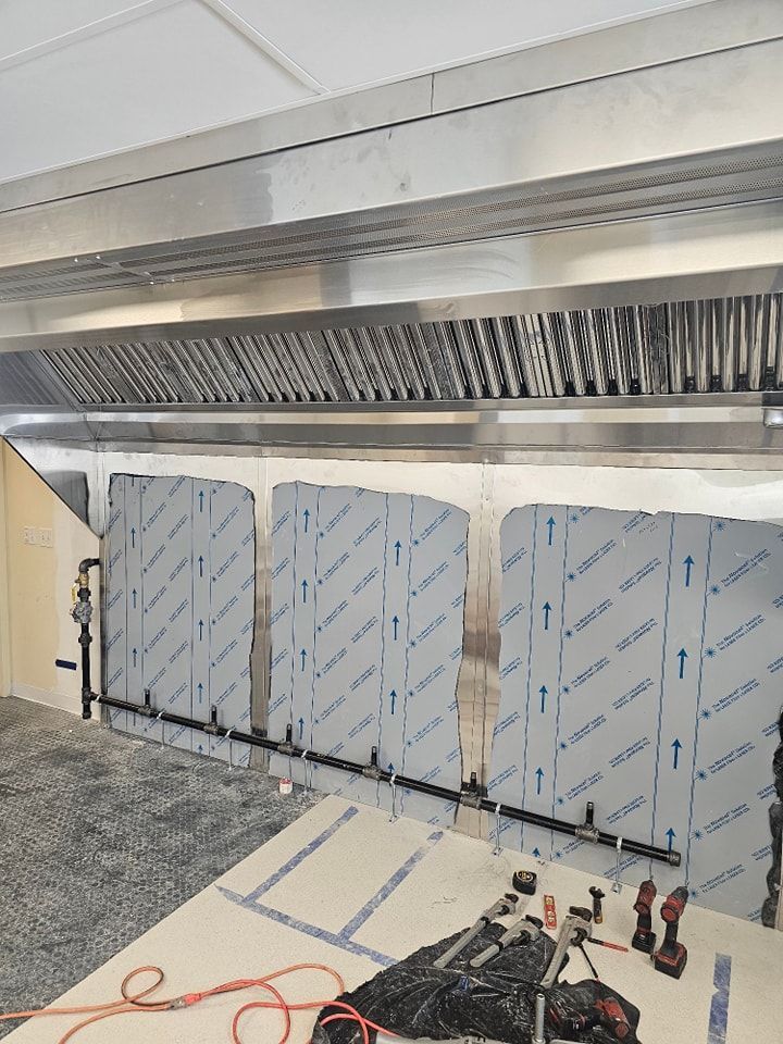 Commercial kitchen under construction: stainless steel hood, metal wall panels, pipes, tools, and a white countertop.