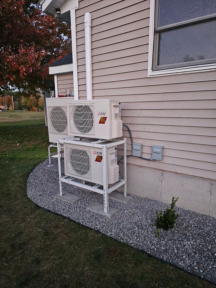 Three outdoor HVAC units mounted on a white rack beside a house with tan siding and a window.