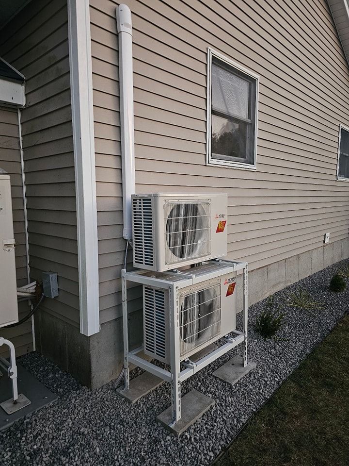 Two white air conditioning units stacked on a metal frame outside a building. White piping runs up the side.