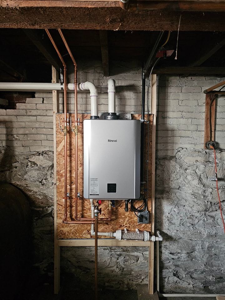Wall-mounted, silver tankless water heater with copper and white pipes in a basement.