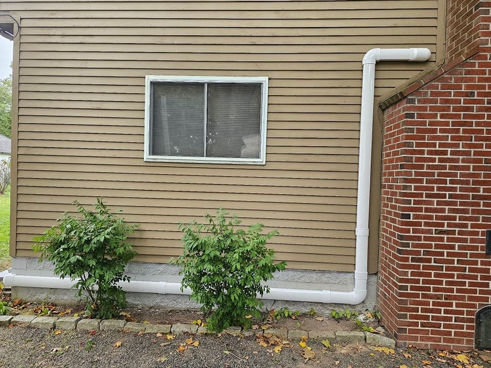 Beige house siding with a white gutter system and a brick chimney, a window, and two bushes.