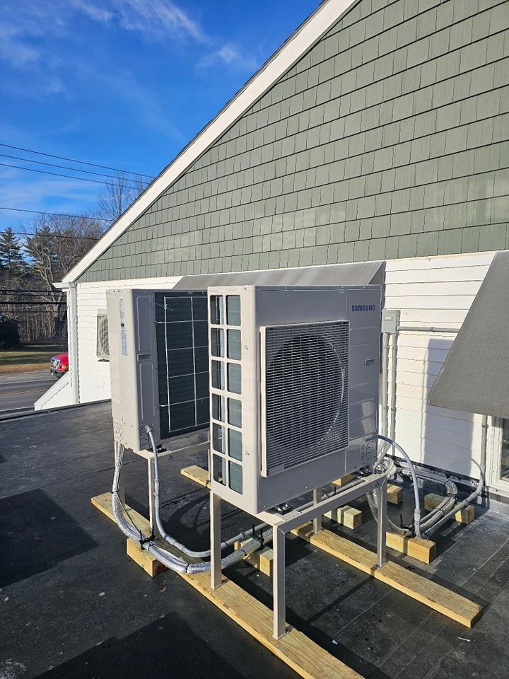 HVAC units mounted on wooden supports on a roof with a green-shingled wall. Blue sky.