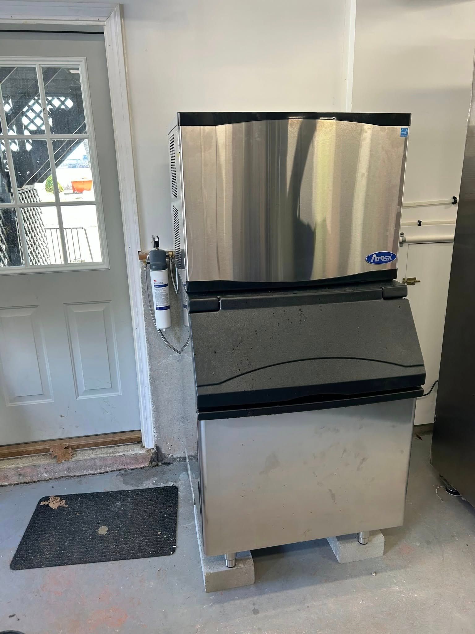 Stainless steel ice machine, next to a door, in a concrete room. Black door mat on the floor.