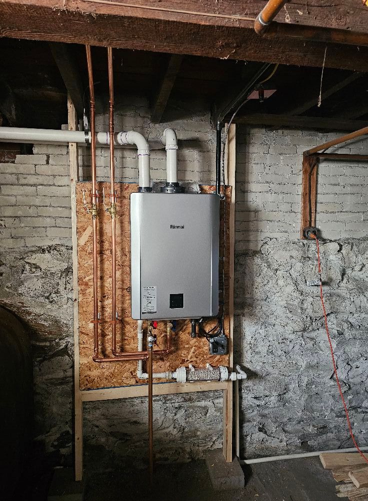 Tankless water heater installed in a basement, mounted on wood. Copper pipes and white vents are visible.