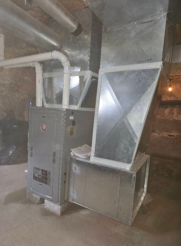 Furnace in a basement, connected to ductwork. Gray metal unit with white pipes against a stone wall.