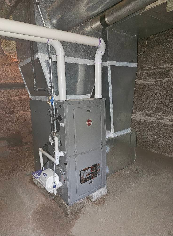 Furnace in a basement. Rectangular metal unit with connected ductwork and PVC pipes, on concrete blocks.