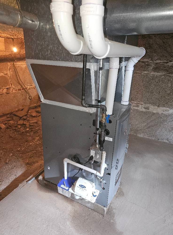 Furnace unit in a basement setting with white PVC pipes, metal ductwork, and grey exterior.