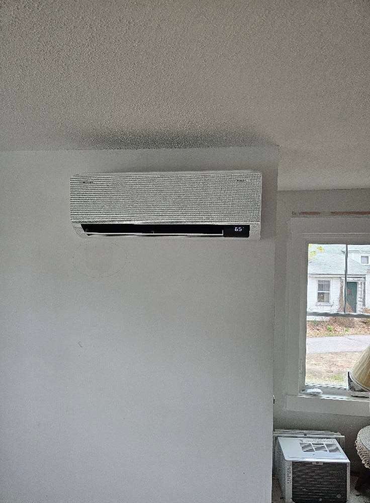 Wall-mounted air conditioner unit in a room with a window, against a white wall and ceiling.